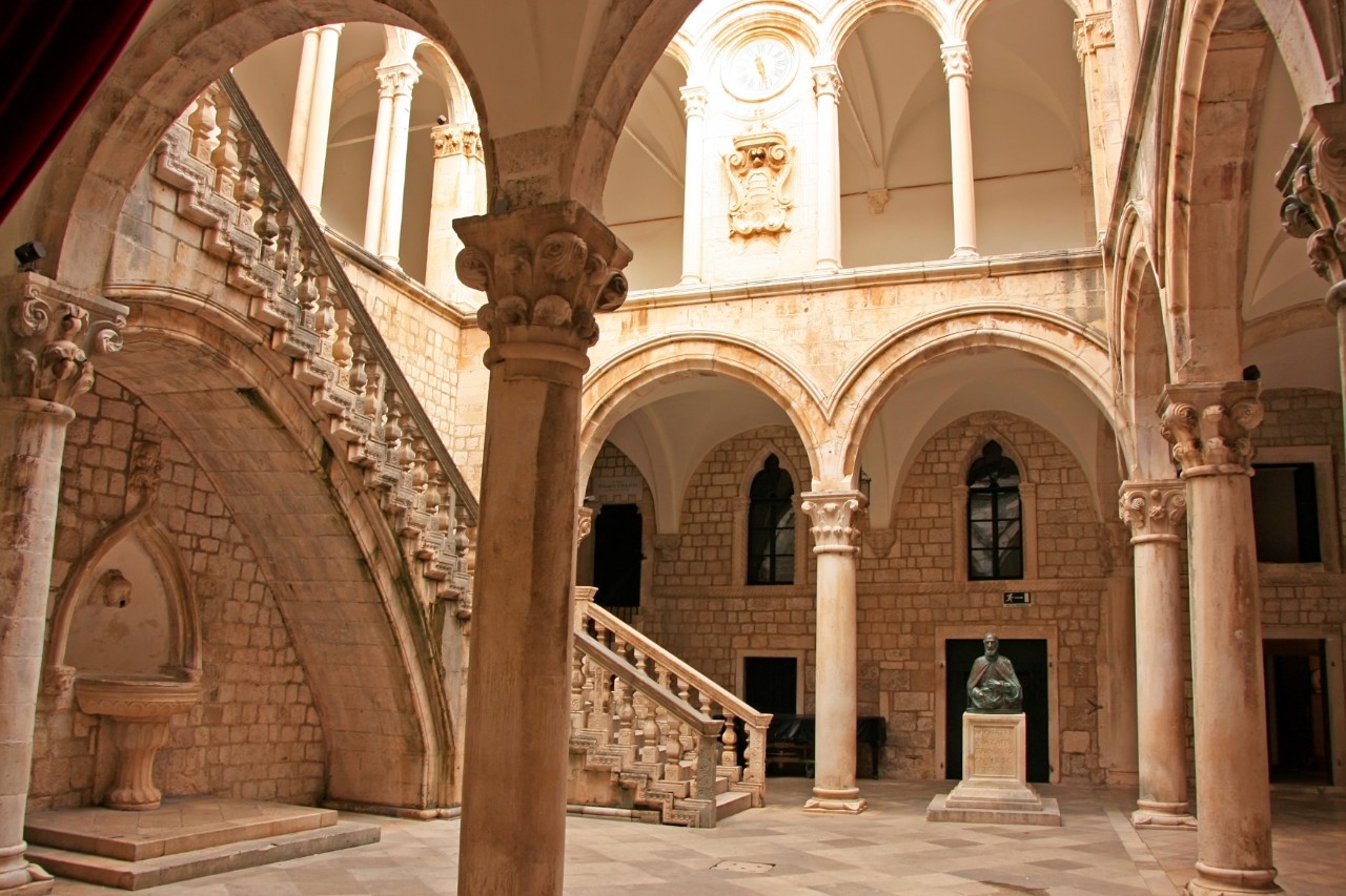The inner courtyard of a light-coloured building, into which the sun is shining from above. Spread over two floors, numerous ornate round arches and columns can be seen and a curved staircase with stone balustrades leads upwards. On the left is a small water basin against the wall.