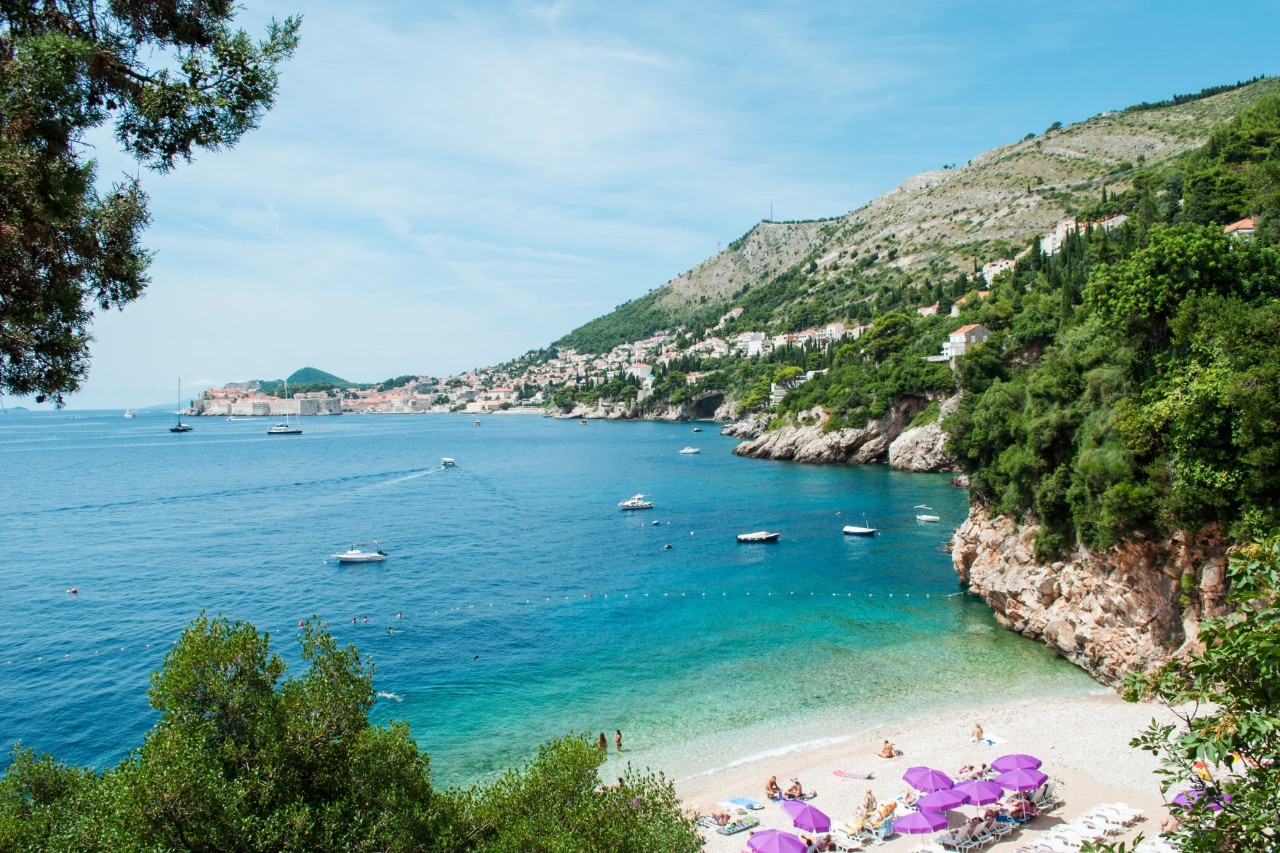 View of a small white bay by the sea, flanked by overgrown rocks on the right and trees on the left. There are white sunbeds, purple parasols and people in swimwear on the beach. A few small boats float on the turquoise water. A city can be seen jutting out into the sea in the background of the picture.