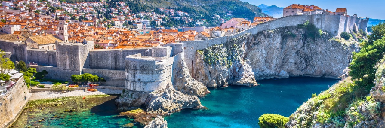 View of a city by the sea, built on a steep cliff and surrounded by a massive city wall. Numerous red roofs can be seen. There are two large fortifications within the city wall. Several small rocks rise out of the turquoise sea in front of the city. A green-covered cliff towers out of the picture on the right.