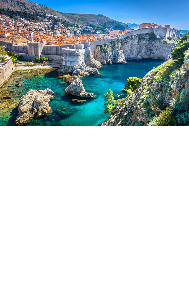 Dubrovnik, built on a steep cliff and surrounded by a massive city wall. Numerous red roofs can be seen. There are two large fortifications within the city wall. Several small rocks rise out of the turquoise sea in front of the city. A green-covered cliff towers out of the picture on the right.