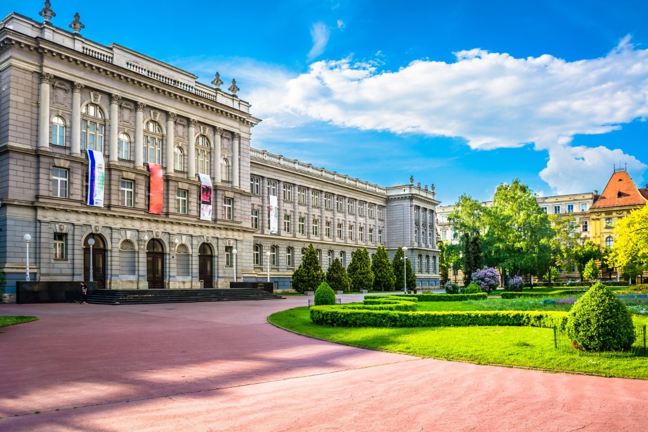 Dreistöckiges Museum im Barock-Stil, davor Grünanlage, im Hintergrund Häuser und Bäume, der Himmel strahlt blau mit Cumuluswolken 