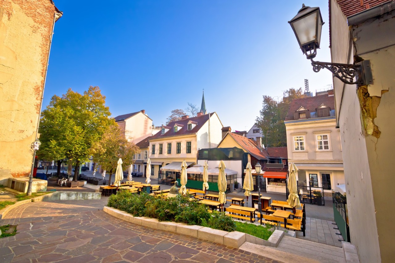Neighbourhood with outdoor restaurant seating, buildings behind, a tree on the far left of the picture 