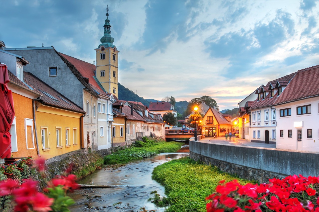 Small river, surrounded by several buildings, a church tower and red flowers 