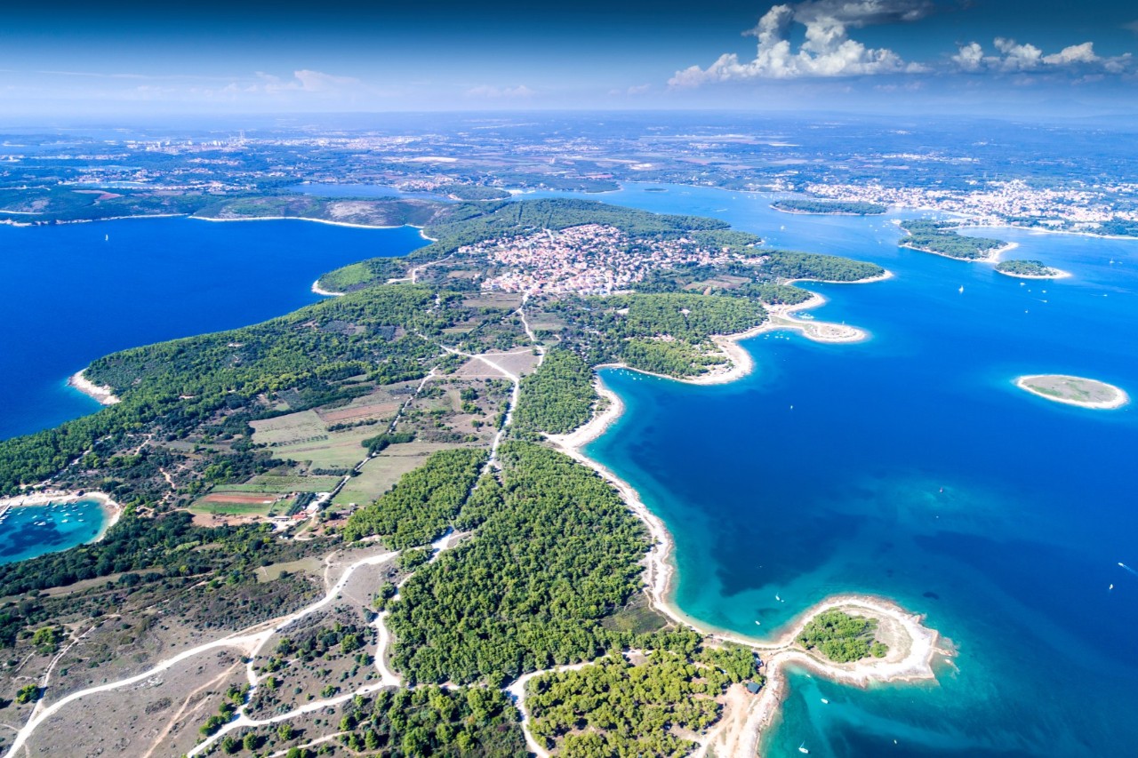 The picture shows an aerial view of a coastal region with numerous bays, clear blue water, and small islands. In the centre is a settlement surrounded by fields, forests, and hiking trails.