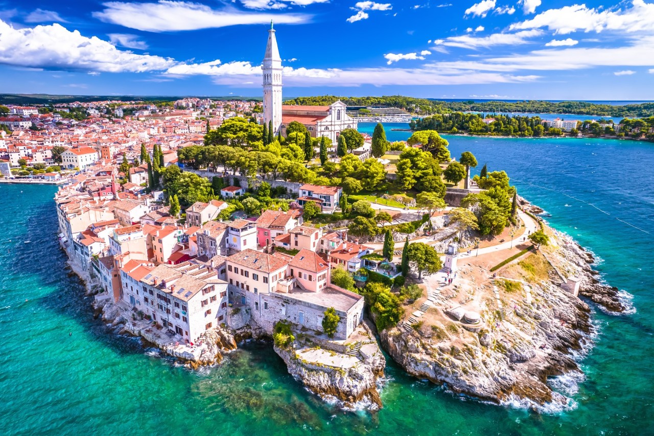 The picture shows a scenic coastal town on a peninsula with historic buildings, narrow streets and a striking church with a high tower. The sea surrounds the settlement, and lush green vegetation is visible.