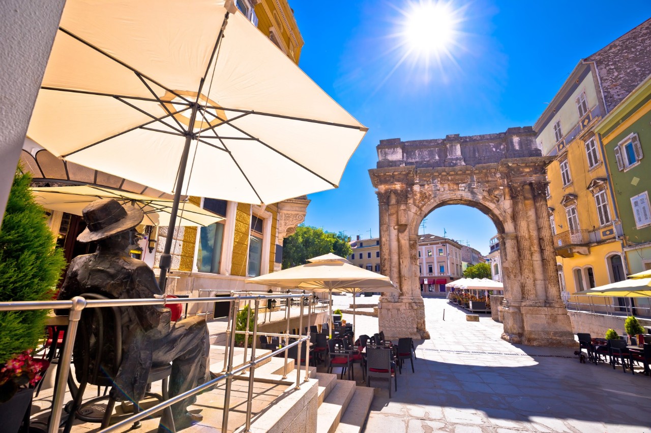 The picture shows a triumphal arch on a cobbled street in bright sunshine. In the foreground, a statue sits on the terrace of a café under parasols. 
