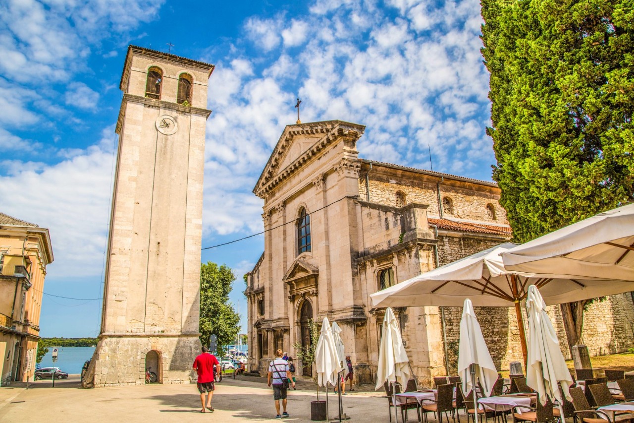 The picture shows a cathedral with a striking, slightly leaning bell tower to its left. In the foreground on the right are empty café tables under parasols, next to which are four closed parasols.