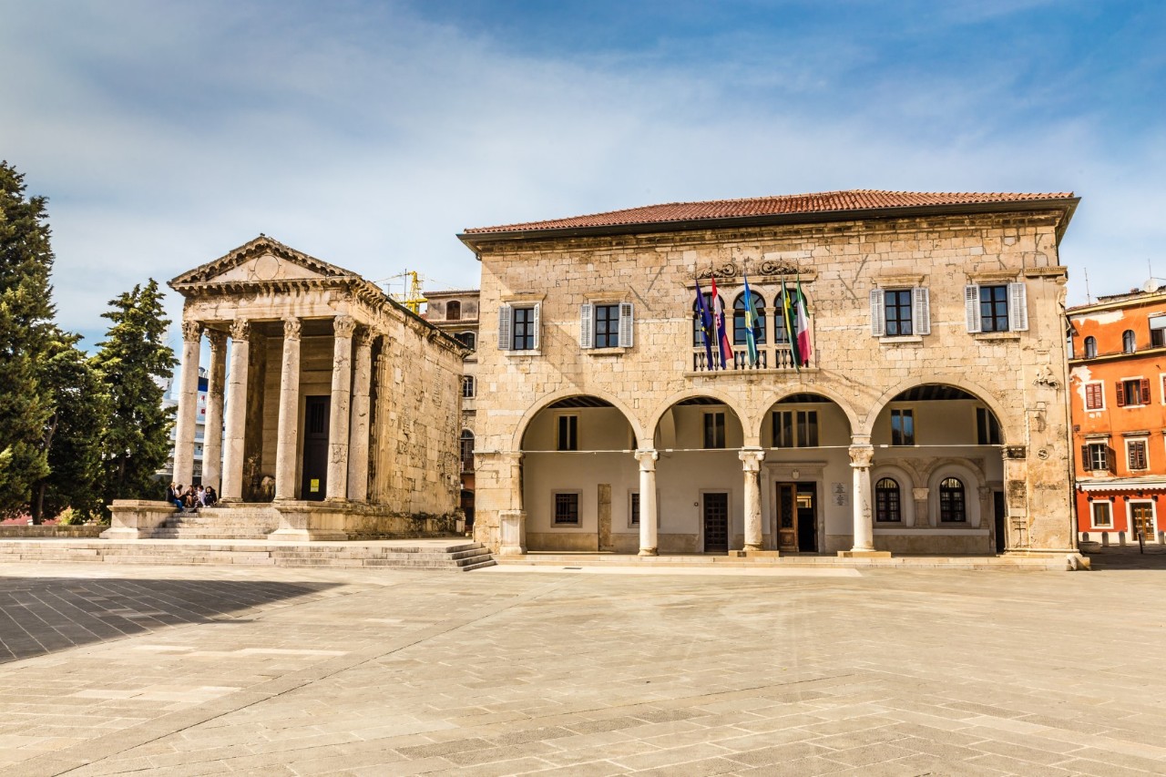 The picture shows a spacious, paved square wi th two striking buildings. On the left is an ancient temple with columns and a tympanum, on the right a building with arcades and flags on the facade.