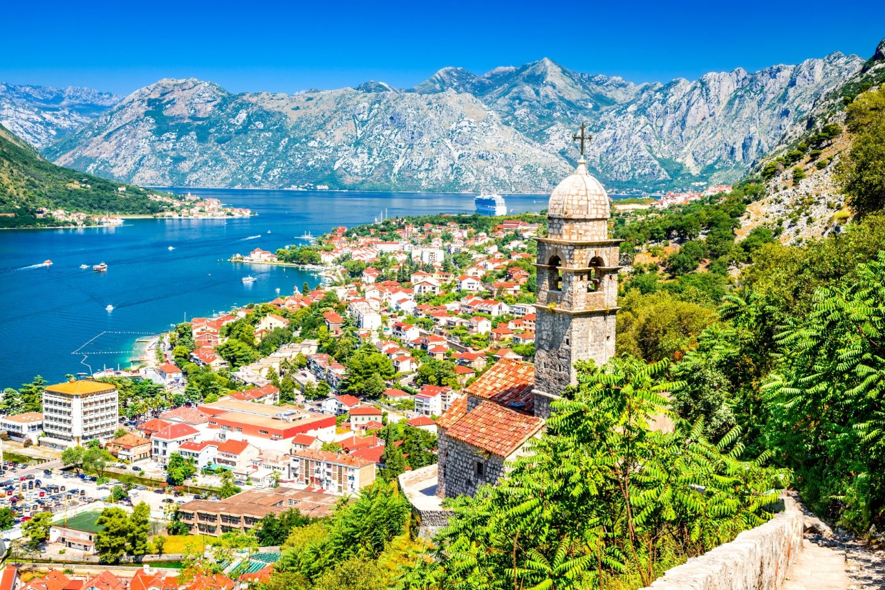 The image shows a coastal town on a bay, surrounded by high mountains and lush green vegetation. In the foreground is an old stone tower with a dome and cross. The houses have red tiled roofs.