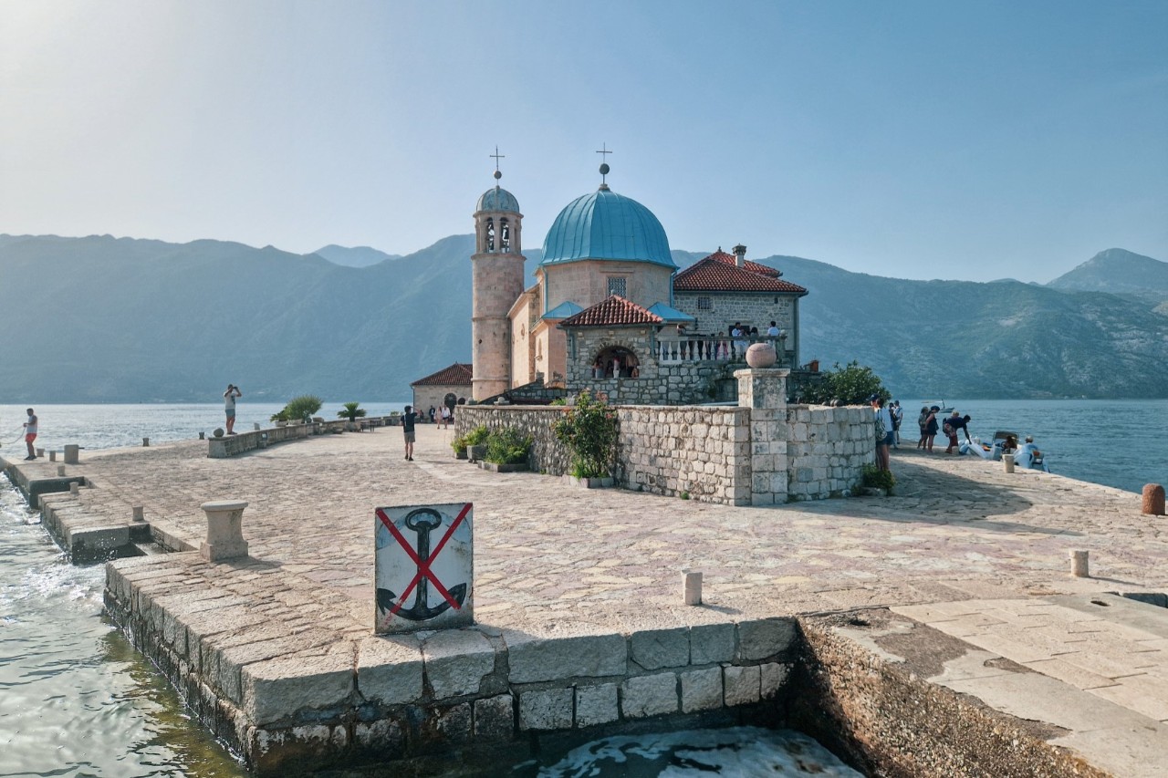 The image shows a church with a blue dome and bell tower on a rocky island. Next to the church, several people are standing by the water. High mountains rise in the background.