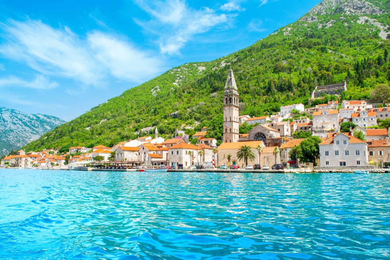 The image shows a Mediterranean coastal town with traditional stone houses and red tiled roofs, situated on the shore of a turquoise blue sea. A striking church tower rises above the town, with green mountains forming the backdrop.