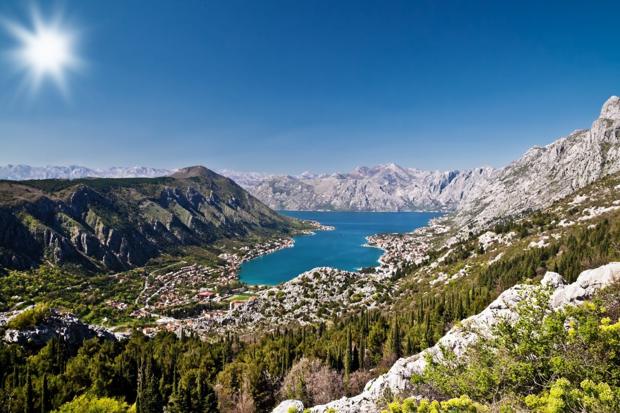 The image shows a deep blue bay surrounded by steep mountains and lush green vegetation. The water winds its way through the landscape, while small, picturesque towns stretch along the coast.