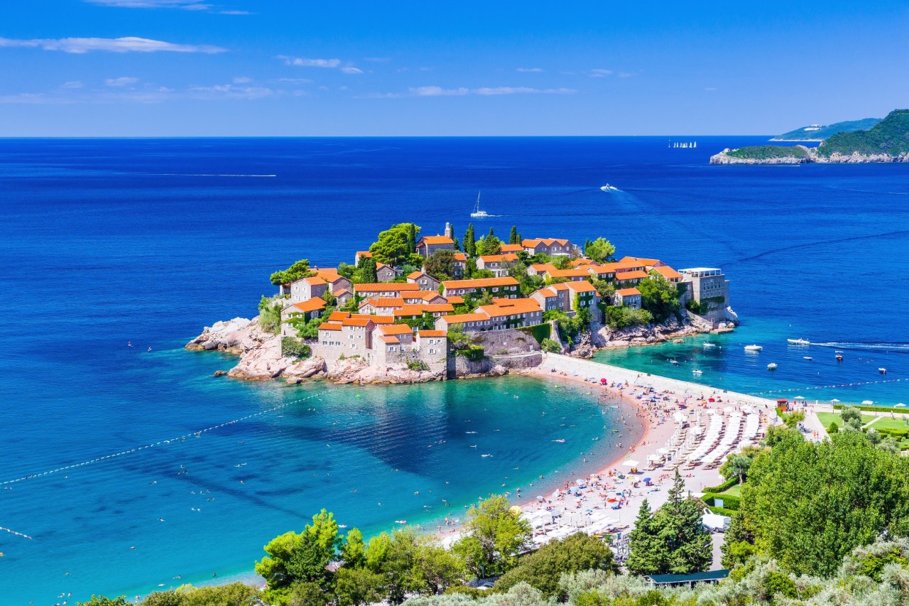 The image shows a small, densely built-up island with historic buildings and red roofs, surrounded by turquoise blue sea. A lively sandy beach leads to the island.