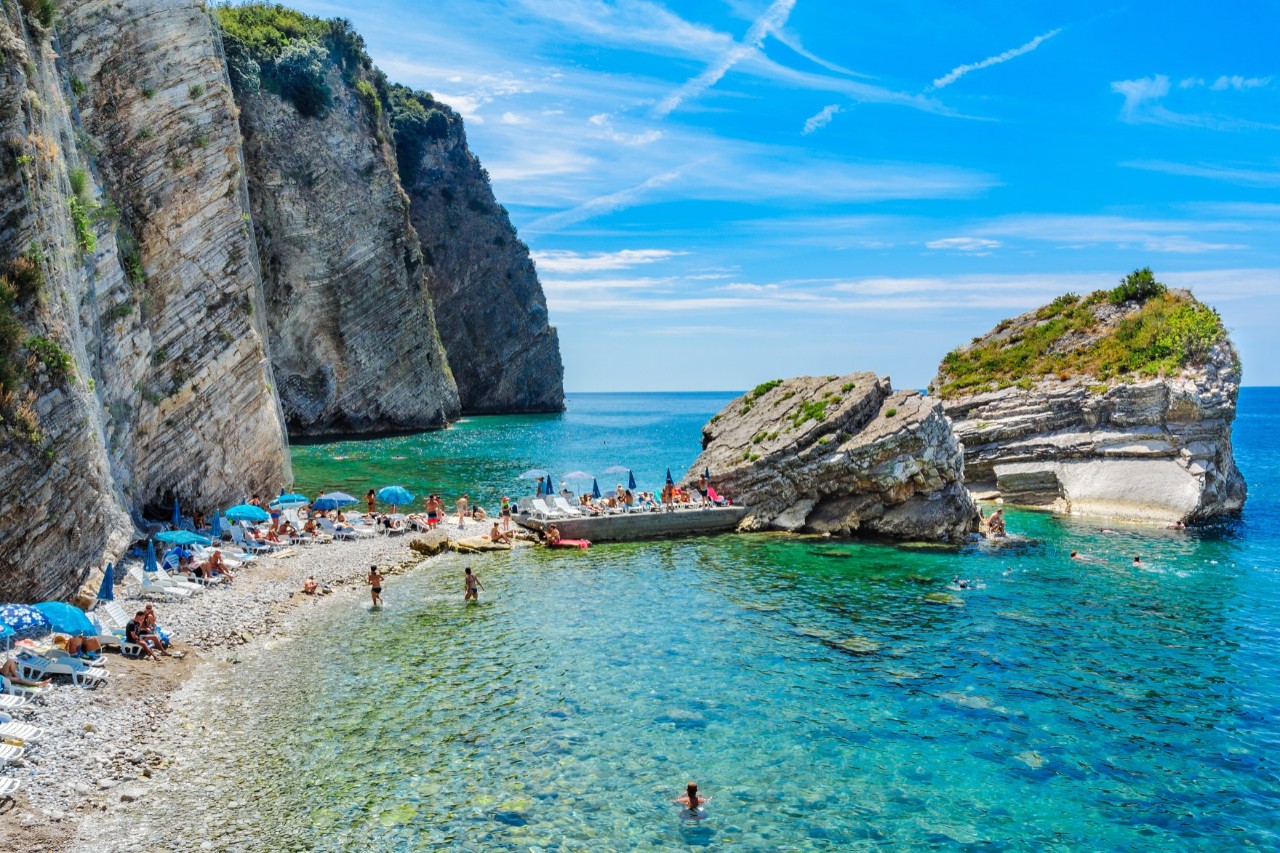 The image shows a very narrow pebble beach with blue parasols and deckchairs, surrounded by steep cliffs and turquoise water. There are people in the water and on the beach.