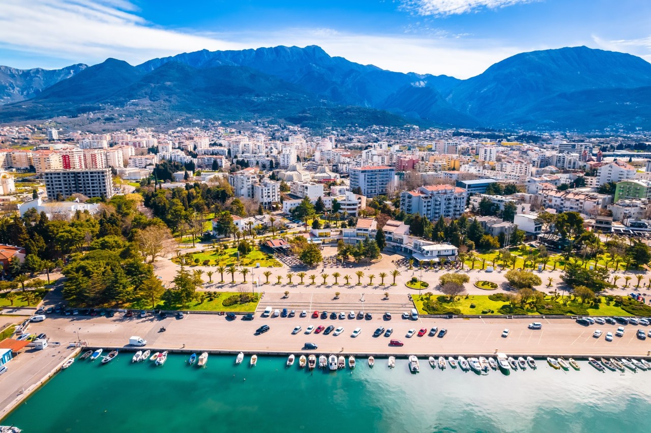 The image shows a coastal town with a promenade where numerous boats are moored. A green park with palm trees stretches out in front of the town. In the background, you can see a dense development of residential buildings and the mountainous hinterland.