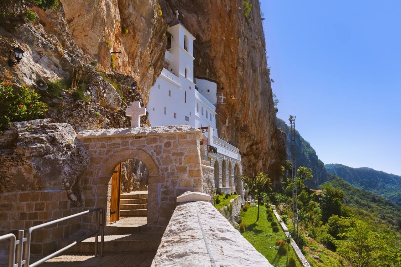 A reddish, steep rock face with a snow-white church built into it. In the foreground, a path separated by a wall leads to the church. An archway made of bricks and a cross above it form the entrance to the monastery grounds.