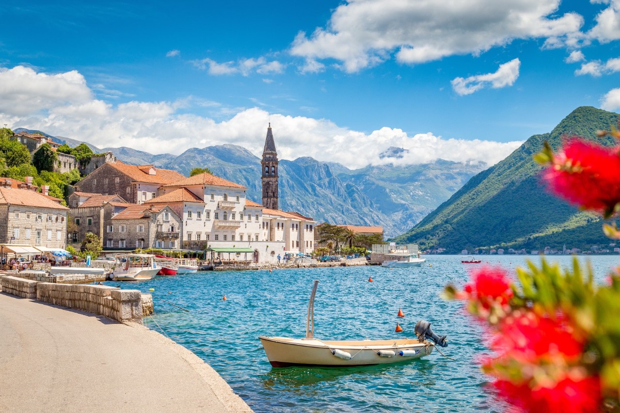 View of a city built right on the shore of a bay. In the foreground, there is a pathway along the water and a boat in the water near the shore. Red flowers poke out of the picture on the right in the foreground. The city consists of numerous bright houses with tiled roofs and a pointed steeple. Mountains can be seen in the background.