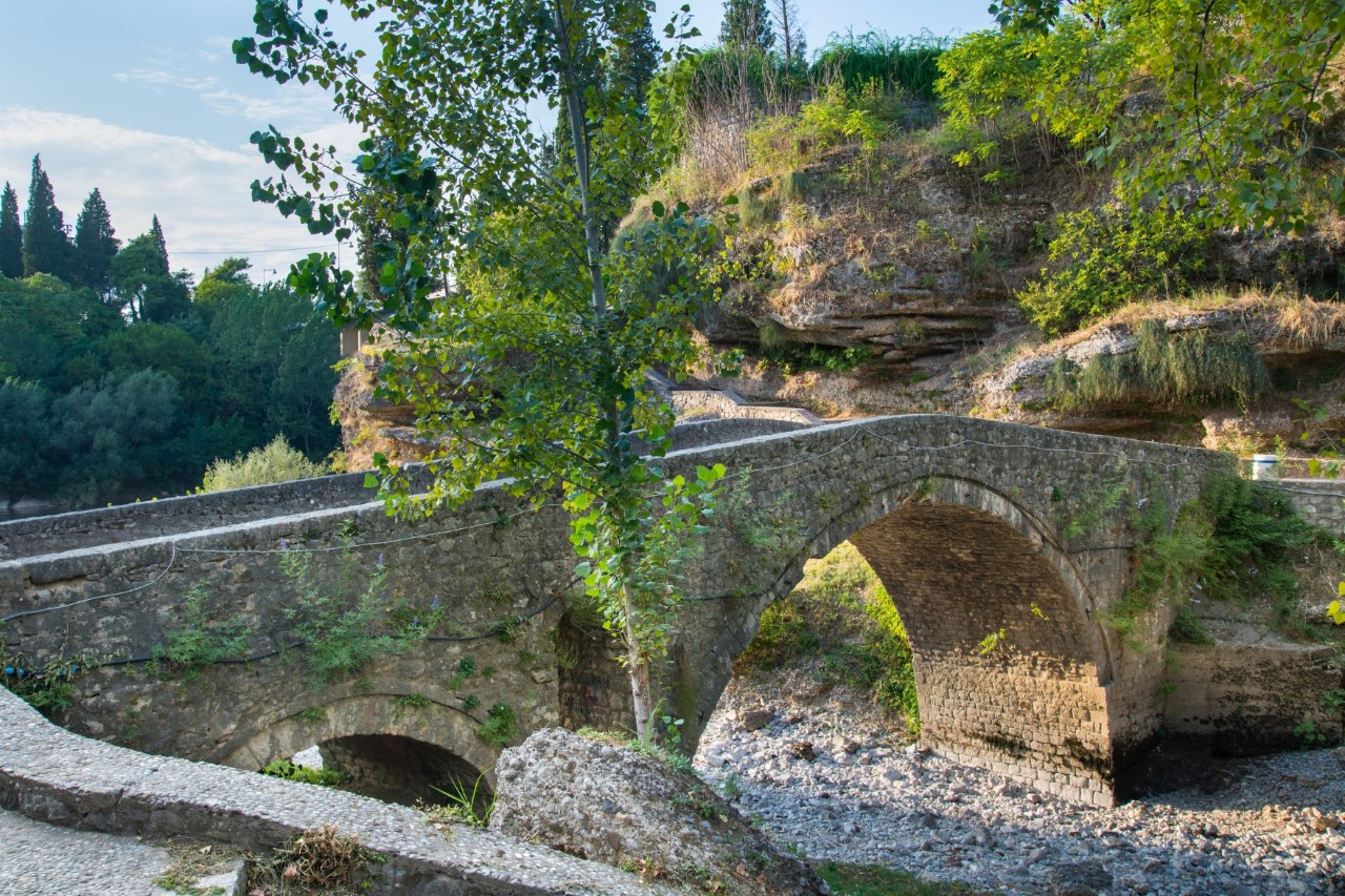 Stone bridge with two arches leading over a dry riverbed. More pieces of a wall and a tree can be seen in the foreground. A hill and forest rise in the background.