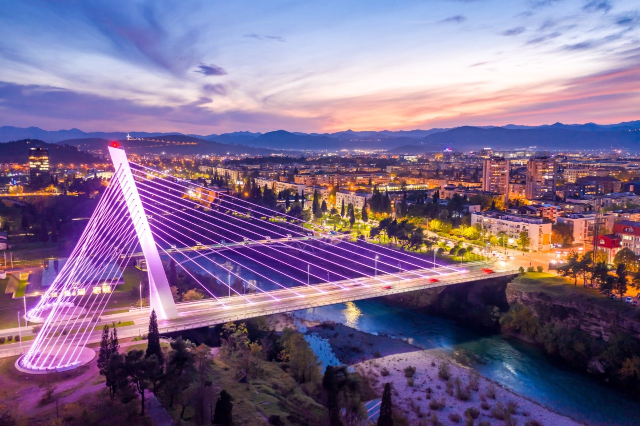 Aerial view of a cable-stayed bridge illuminated in pink in the evening light, connecting two riverbanks in a city. The city's houses and streets are lit up on both riverbanks; the evening sky is bathed in many colours.