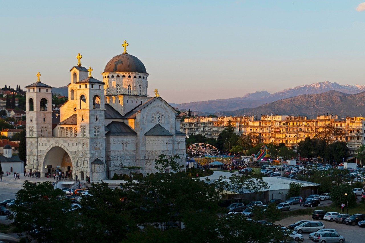 Large, bright cathedral with several towers and a round, dome-shaped main tower. Each spire is topped with a cross. The roofs of the towers are grey. A large, arched main entrance is at the front. There is a bright square around the cathedral, with houses and hills glowing in the evening sun in the background.