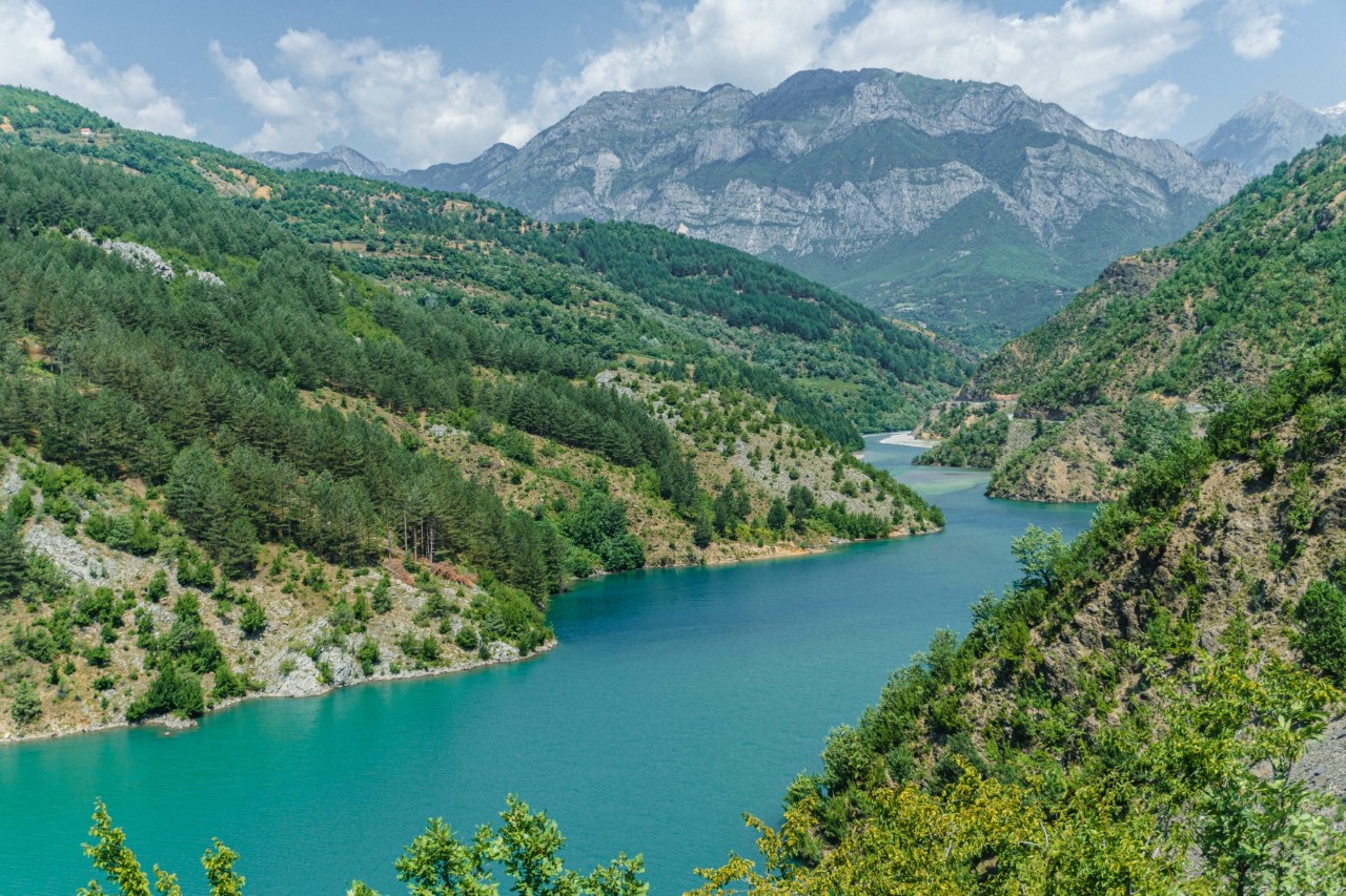 Lake surrounded by mountains and green hills. The turquoise water winds its way through the forested hills. Rocky mountains rise in the background.
