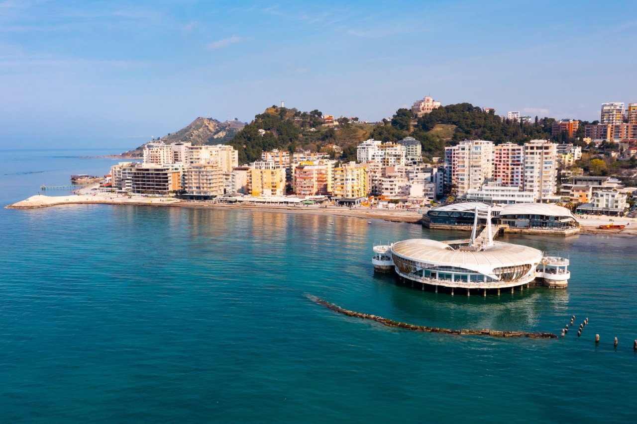 Coastal town with modern high-rise buildings and a striking round building on stilts in the sea. Green hills can be seen in the background.