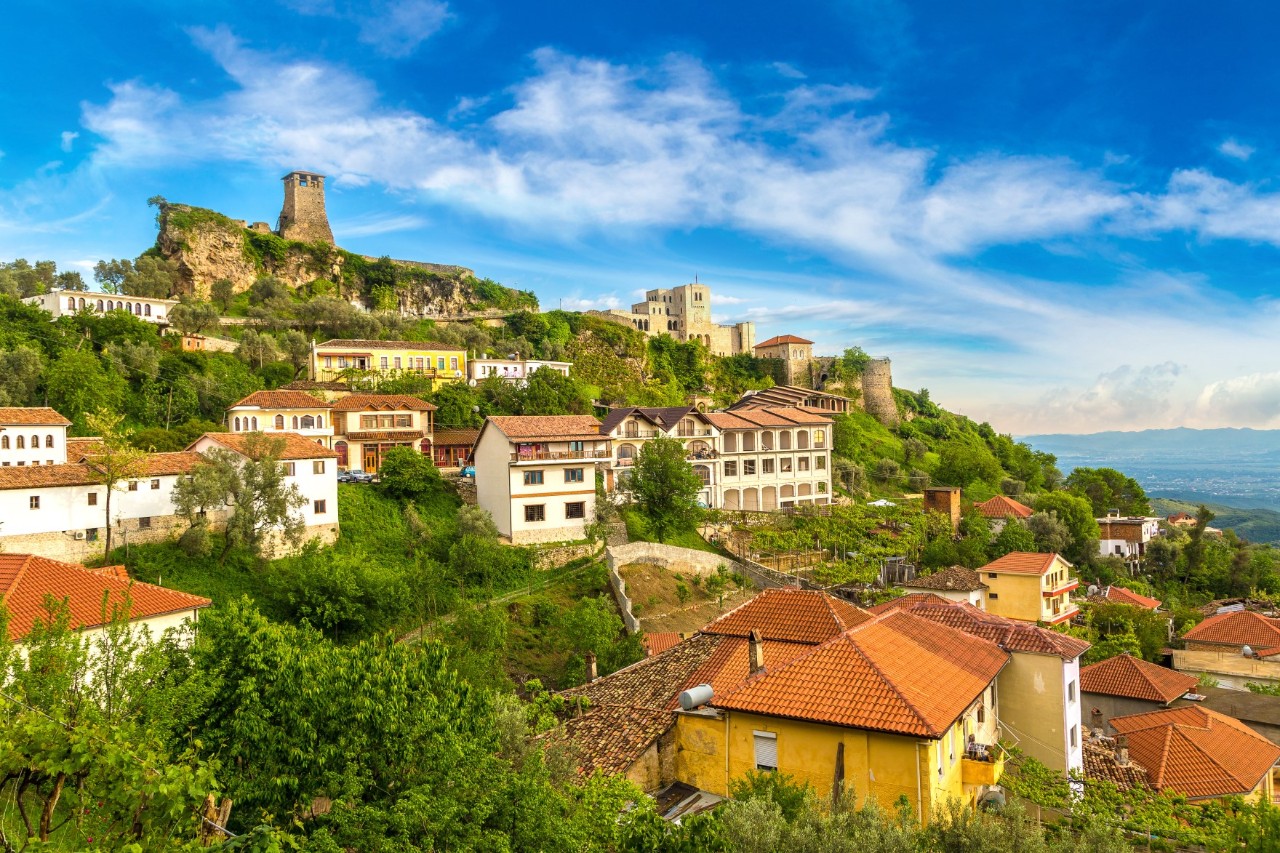 Village stretching up a green hillside. At the very top of the hill stands an old castle. The traditional houses are white or yellow and have red tiled roofs.