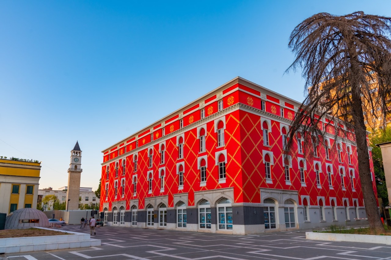 Striking red building with a yellow diamond pattern on the facade. In the background, there is a historic clock tower. The square in front of the building is spacious and has a pattern on the ground. There is a tree on the right side of the picture. 