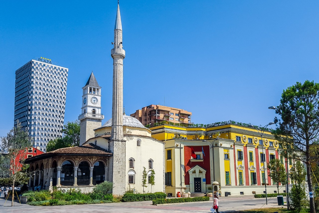Mosque with a minaret and a clock tower. The mosque has a vaulted entrance with columns. Next to it is a yellow-and-red building. In the background, there is a modern high-rise.