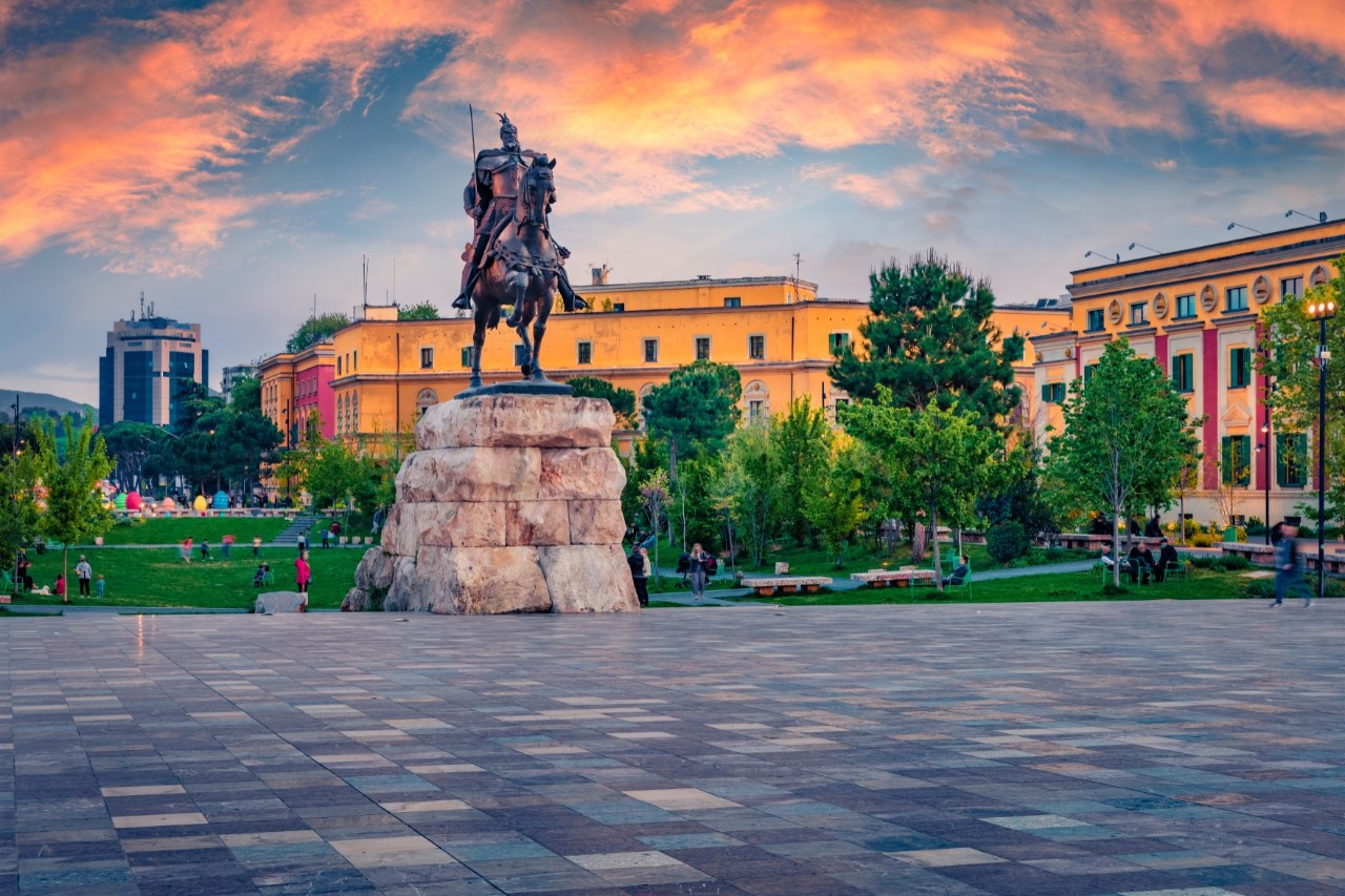 Equestrian statue in a large square. In the background, there are colorful buildings and a green park with trees and walking paths. The sky is bathed in warm colors.