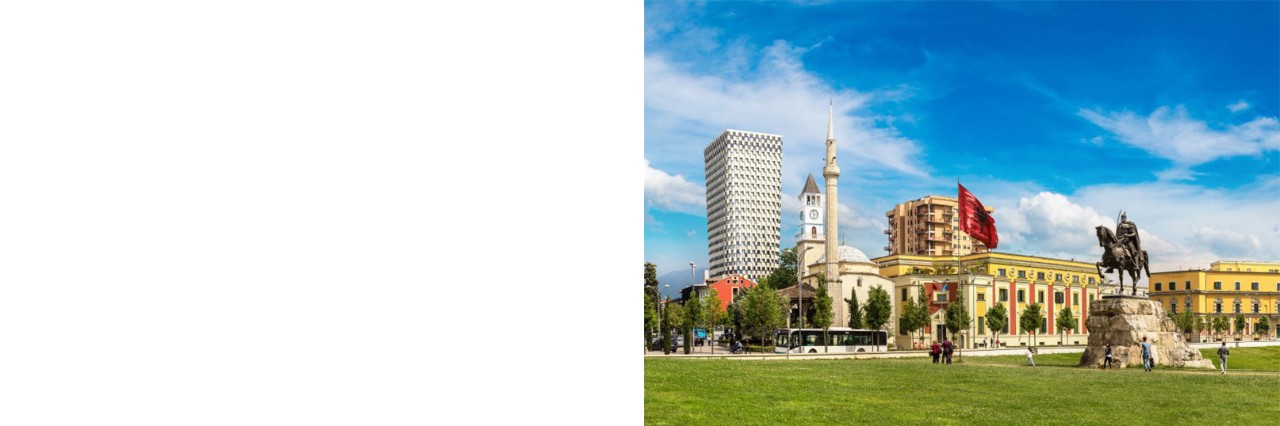 Large meadow with an equestrian monument in the middle. Several buildings can be seen in the background, including a mosque with a minaret, a high-rise building and a clock tower as well as several elongated, multi-storey yellow houses. A red flag flies on a pole. The sky is slightly cloudy. 