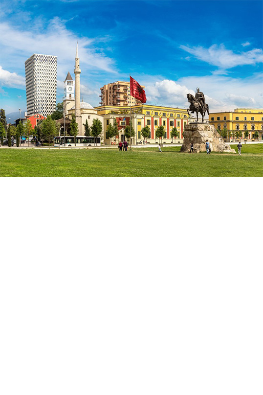 Large meadow with an equestrian monument in the middle. Several buildings can be seen in the background, including a mosque with a minaret, a high-rise building and a clock tower as well as several elongated, multi-storey yellow houses. A red flag flies on a pole. The sky is slightly cloudy. 