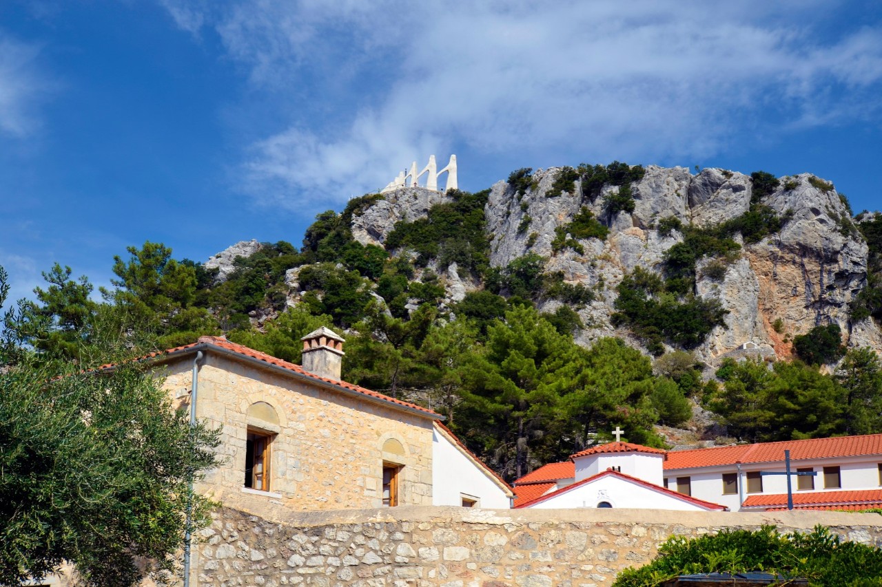 A mountainous landscape with traditional stone houses in the foreground. In the background, a rock covered in green vegetation rises up, topped by a striking monument with white figures.