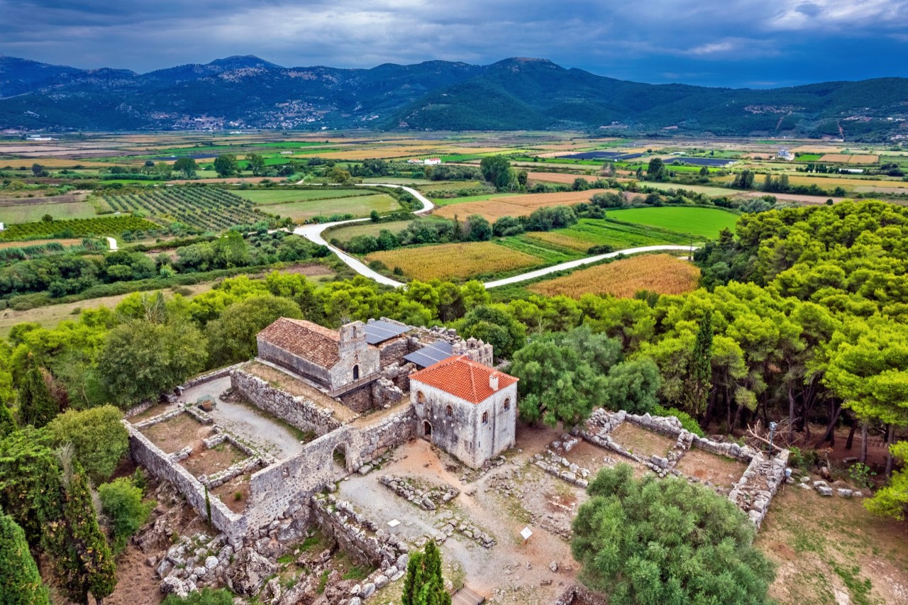 The image shows ruins with stone walls and two preserved buildings, one with a light red roof. The complex is surrounded by green trees and fields, with a mountainous landscape in the background. The sky is dark blue.