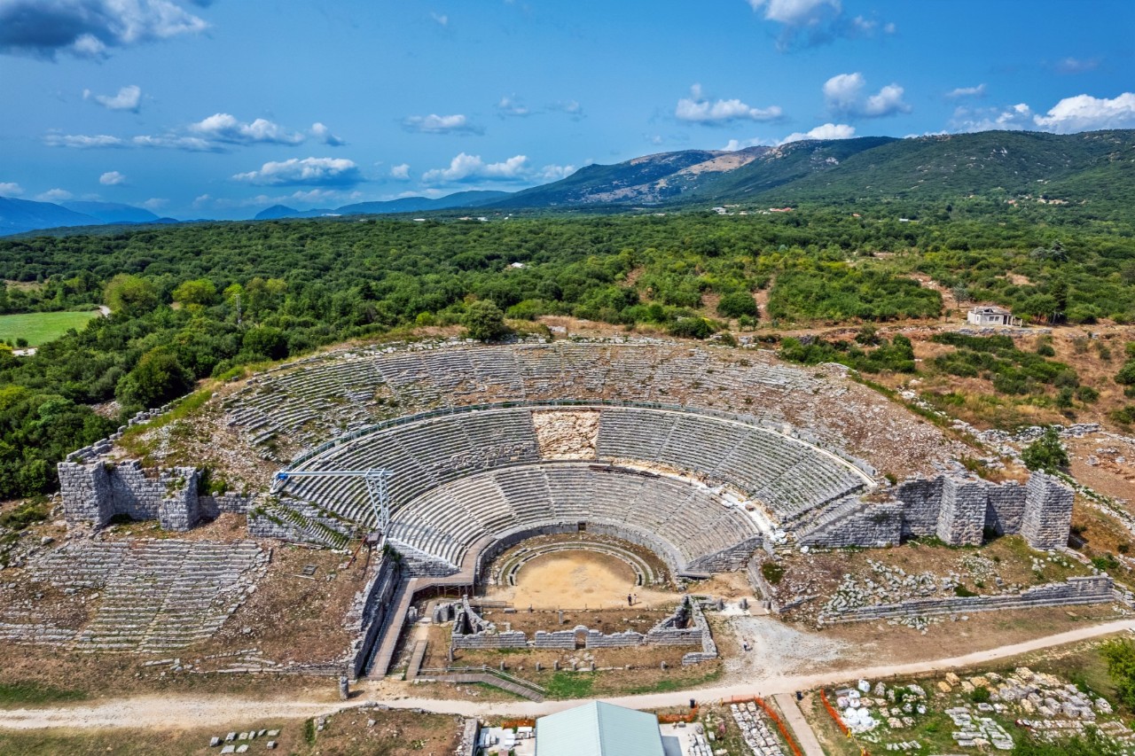 View from above of an ancient amphitheatre surrounded by green countryside with hills in the background. The semi-circular stone seating rows are well preserved, while some of the walls are in ruins.