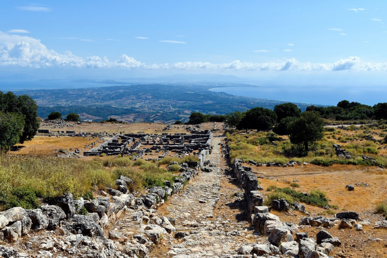 The image shows an ancient ruin on a hill overlooking a vast landscape and the sea. An old paved path leads through the remains of walls and buildings, surrounded by dry grass and scattered trees.