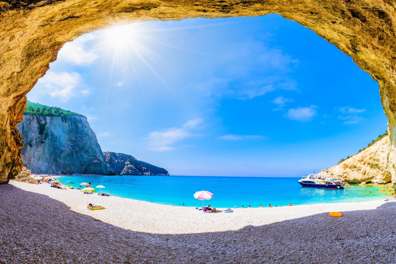 View through a cave onto a flat beach and the turquoise to blue sea. There are a few people on the beach, a boat is moored off the coast, and mountains surround the bay.