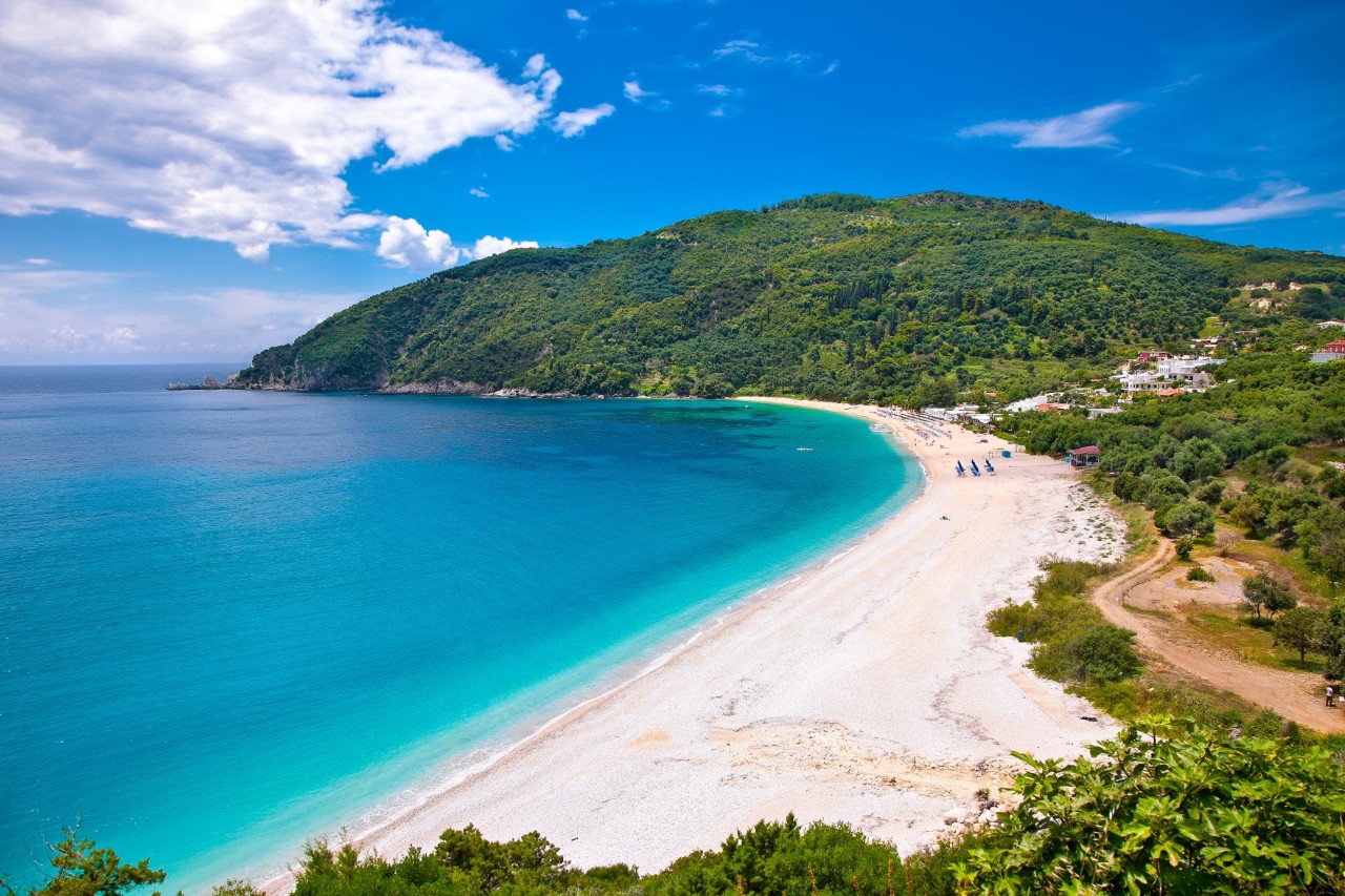 The image shows a long, white sandy beach along the turquoise blue sea. The surroundings are characterised by densely vegetated mountains and hills.