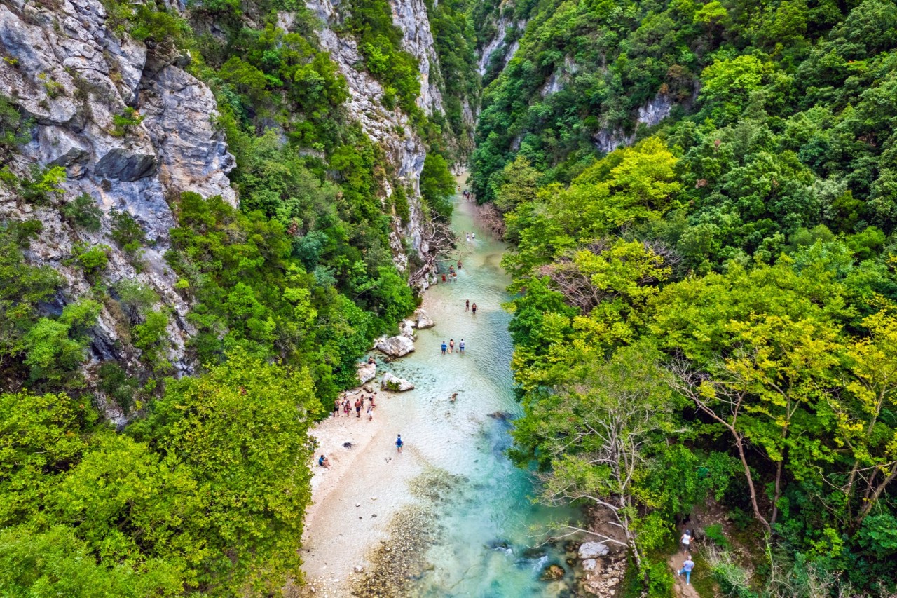 Bird-eye view of a river landscape with people in a gorge with turquoise water. The surroundings consist of steep cliffs and dense greenery.