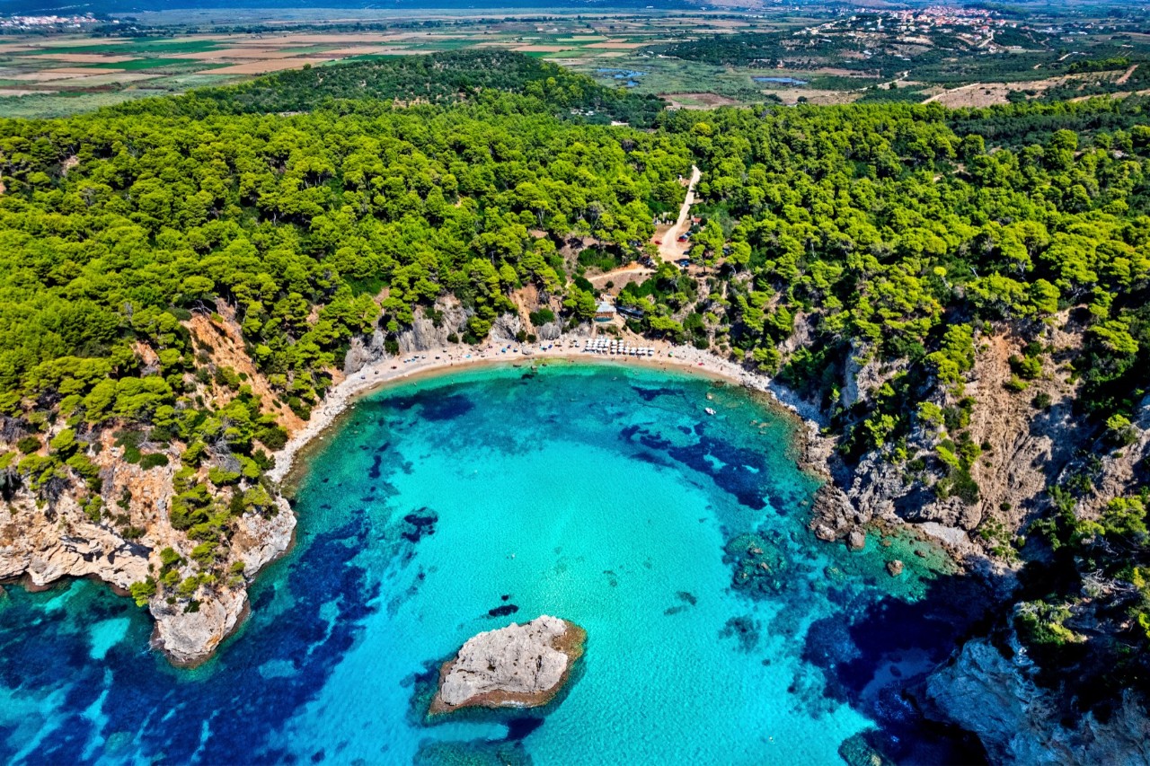 The image shows a semi-circular bay with turquoise blue water, surrounded by densely wooded cliffs and a sandy beach. A rock rises out of the water in the centre.