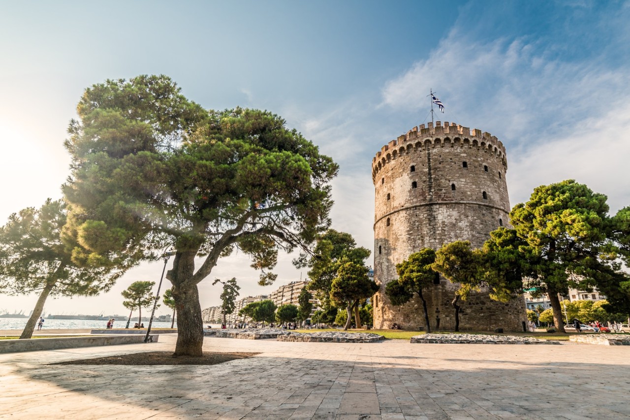 The picture shows a flag flying atop a huge, cylindrical stone structure surrounded by lush greenery and trees. Modern buildings and the waterfront can be seen in the background.