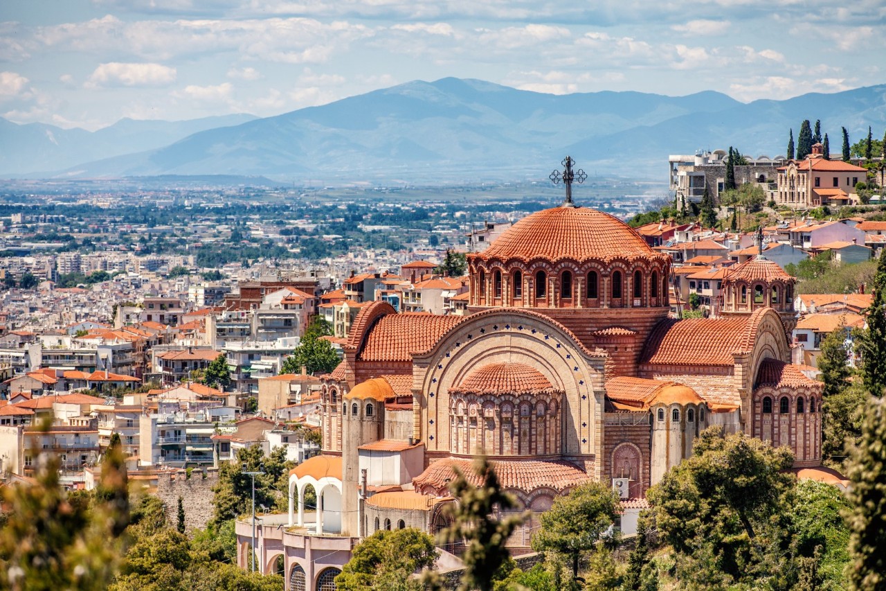 The picture shows an impressive Orthodox church in the typical Byzantine design with a large dome, several smaller towers and red-tiled roofs. Also captured are the densely built-up city in the background and a mountain range on the horizon.