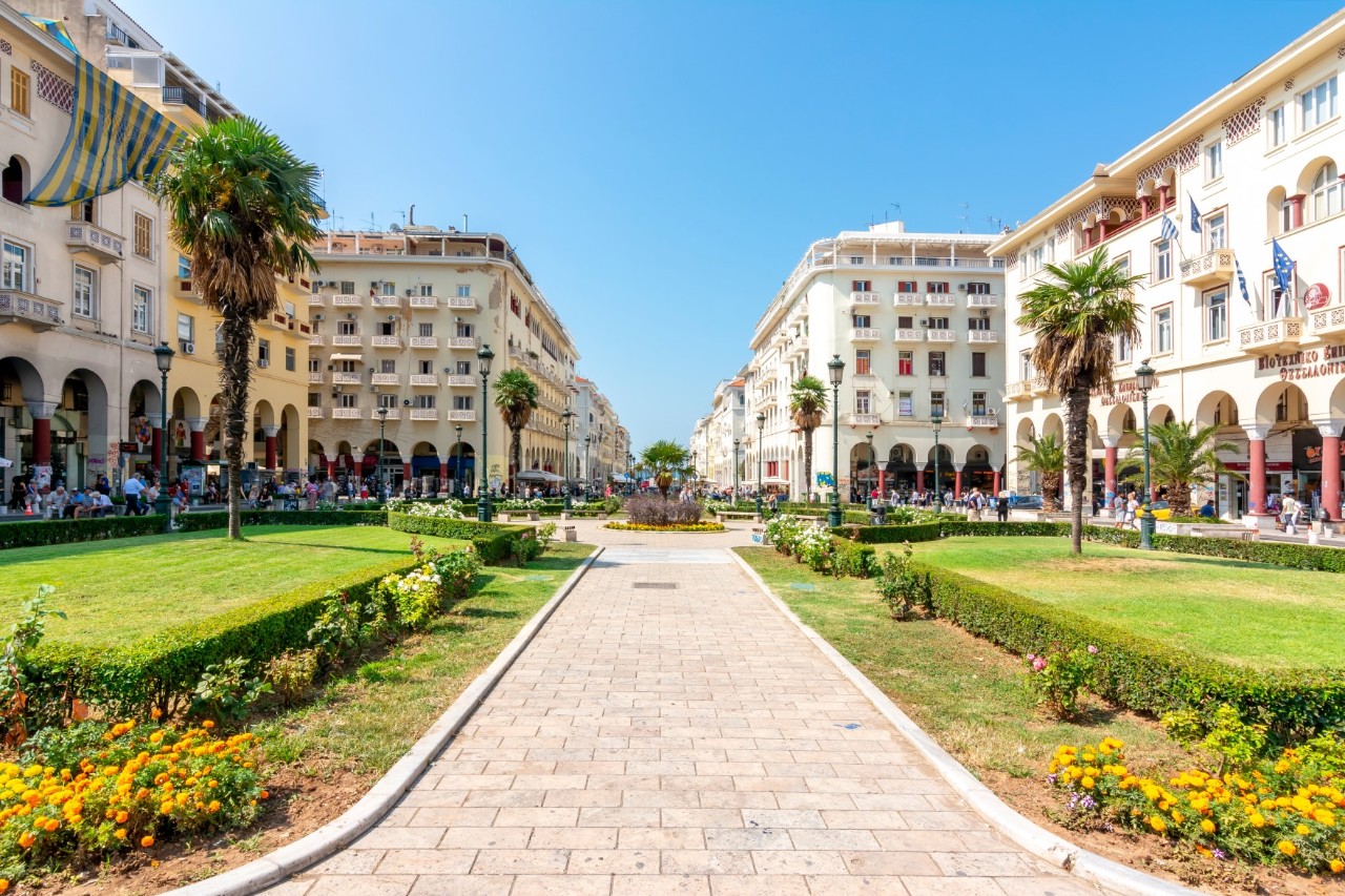 The picture shows a large, open square flanked by elegant, white buildings with arcades. In the middle of the square, there are well-kept green areas with flowerbeds, palm trees and walkways. People can be seen in the background milling around the square. The sky is clear and blue.