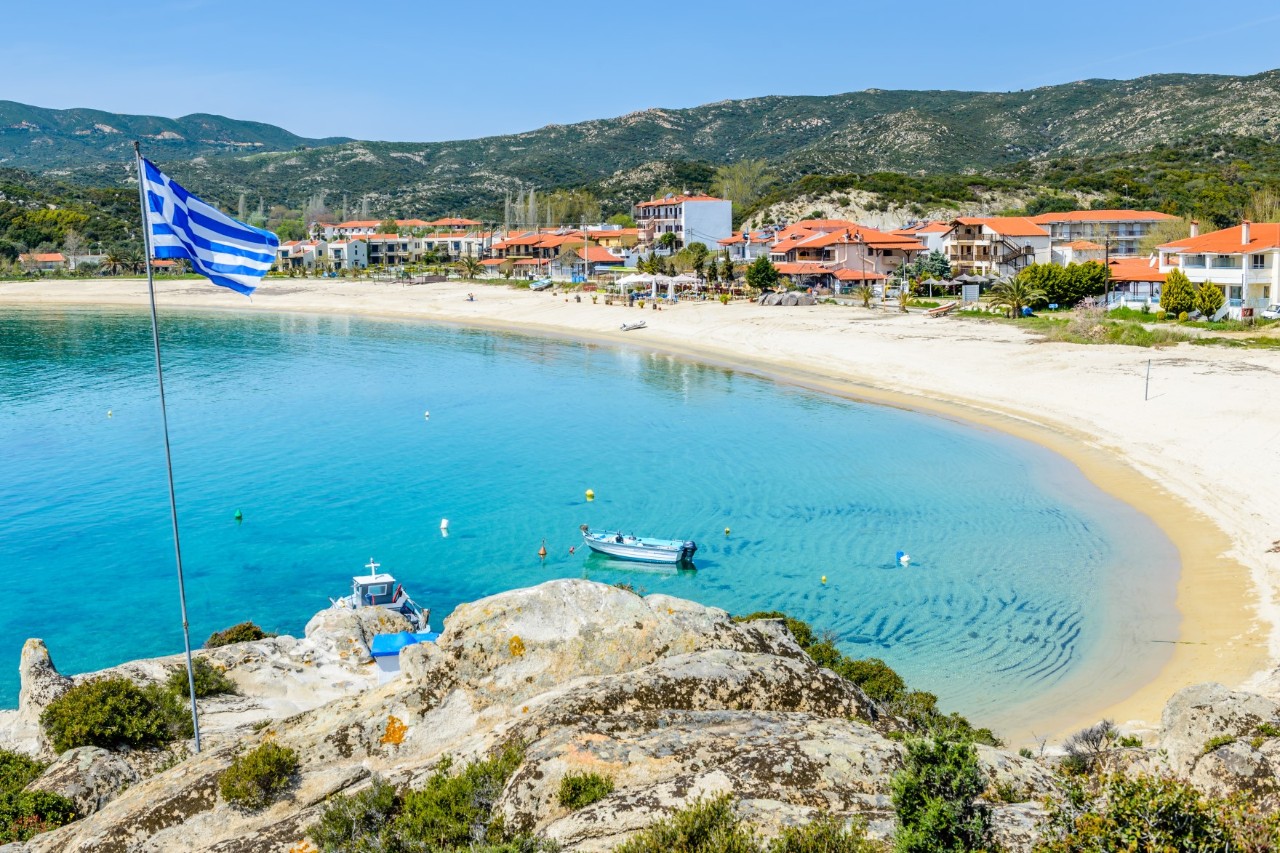 The picture shows a quiet bay with a small, picturesque village. Several houses with red-tiled roofs line the coast next to the clear water where a few boats are moored. A Greek flag flies in the foreground and rocky sections of coast can be seen.