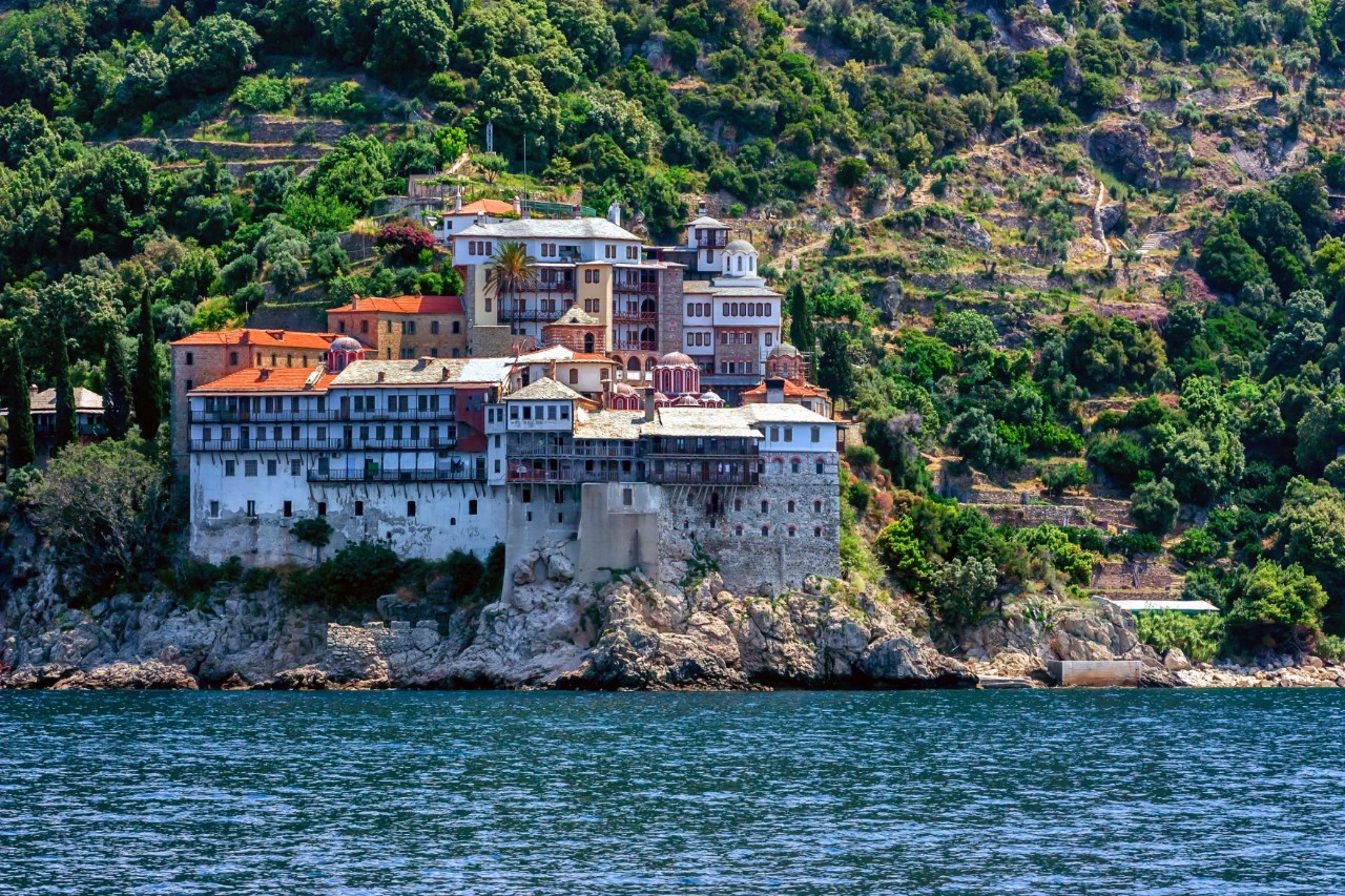 The picture shows an impressive monastery built on a steep cliff directly by the sea. The monastery buildings are tightly nestled together, seemingly growing out of the rock. The monastery is surrounded by lush green vegetation and the sea in the foreground is calm and deep blue.