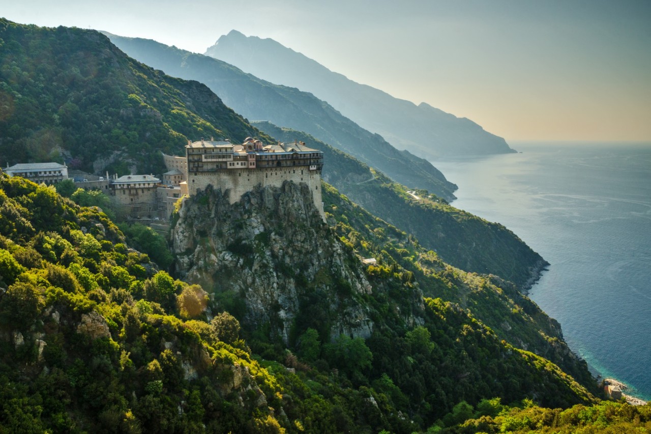 The photo shows an imposing monastery built on a steep rock high above the sea. It is surrounded by densely forested hills. The coastline extends in the background and on the right-hand side of the picture.