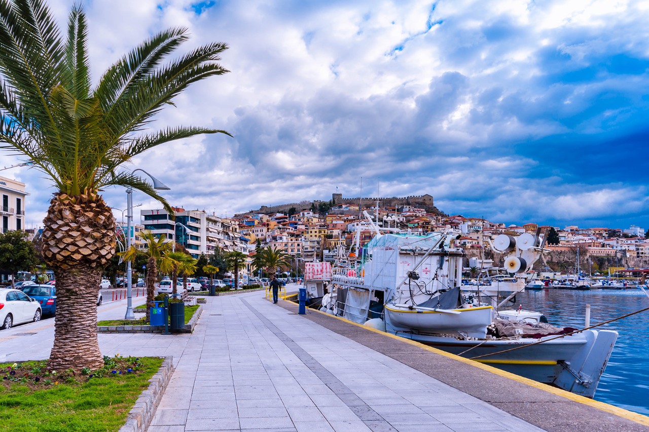 Port promenade with small boats in the water. Palm trees line the shore. The city and its fortress rise in the background.