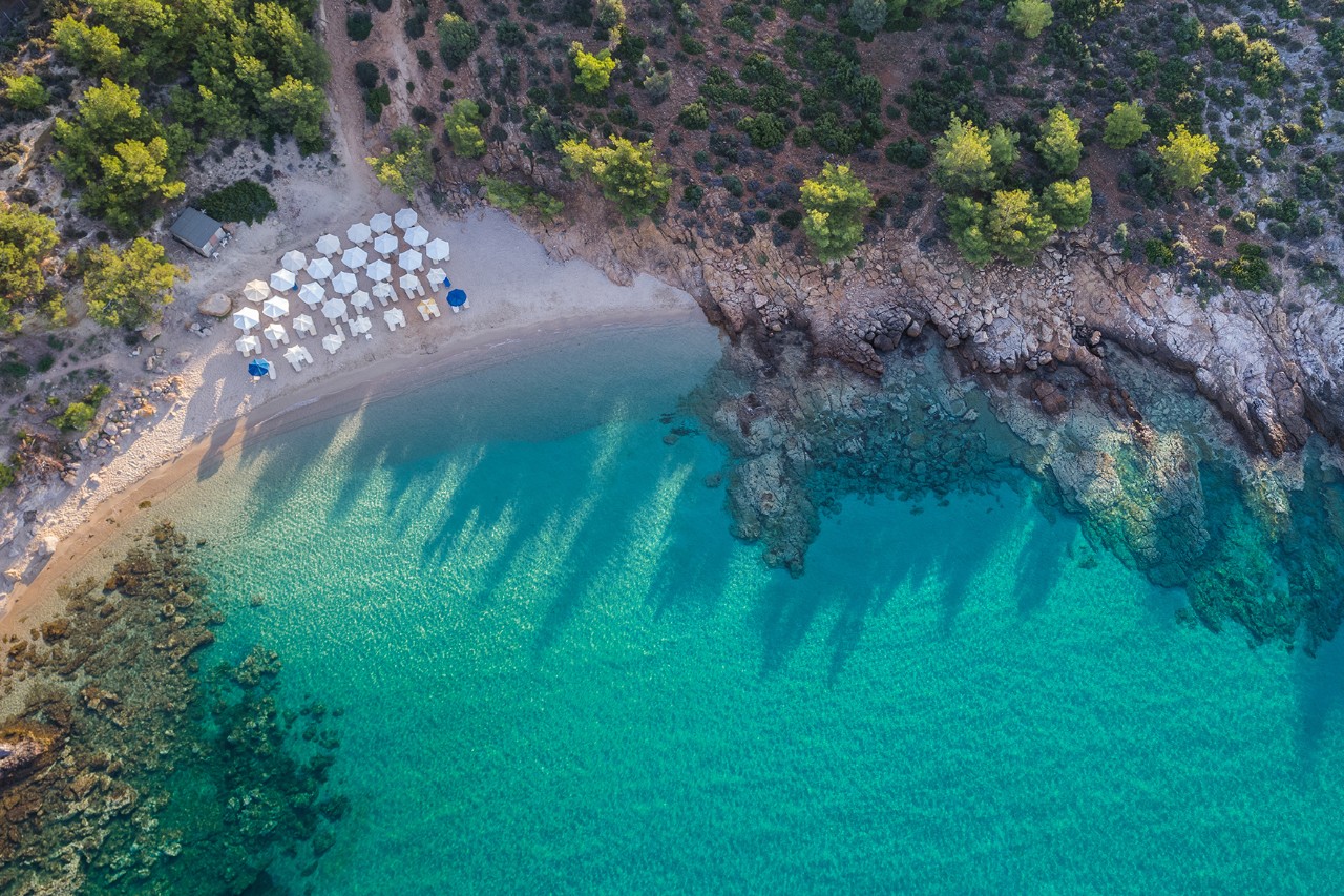 Aerial view of a bright, sandy beach with turquoise, clear sea. Parasols and sun loungers are arranged on the shore, behind which are green slopes and rocky coastlines.