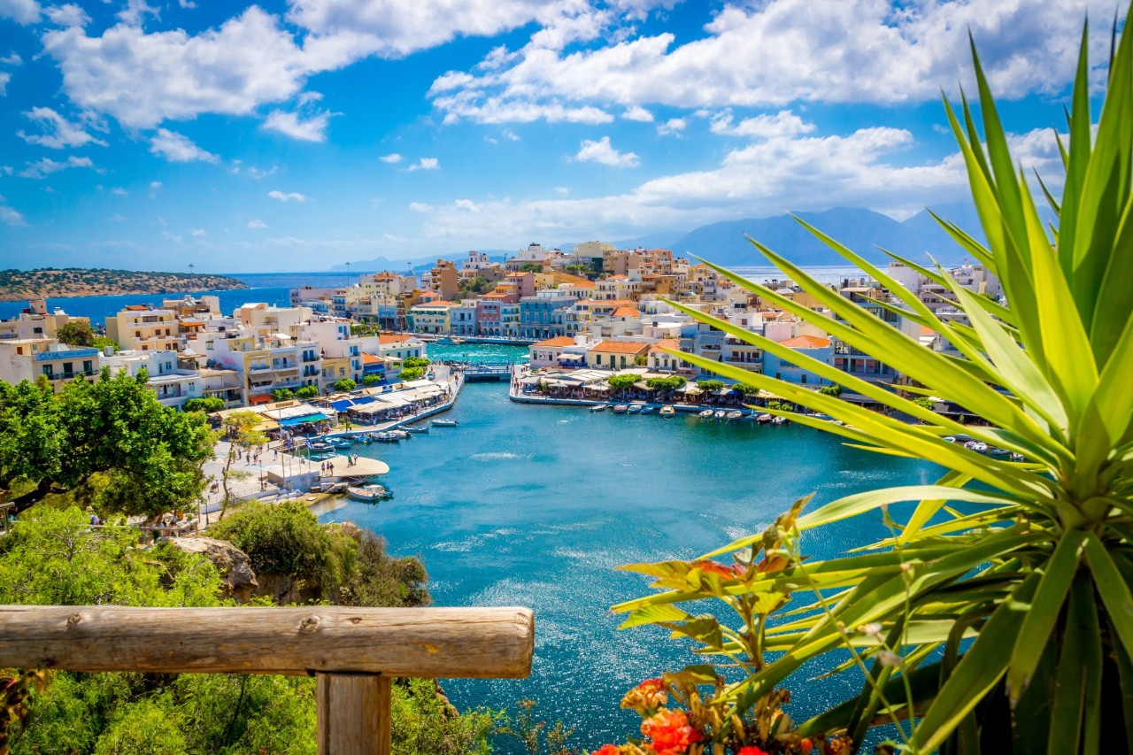 A turquoise lake. It is surrounded by a harbour with small boats and surrounding houses. The sea can be seen on the horizon. The sky is blue with smaller cumulus clouds. Green trees can be seen in the foreground on the left, and a dragon tree extends into the picture on the right.