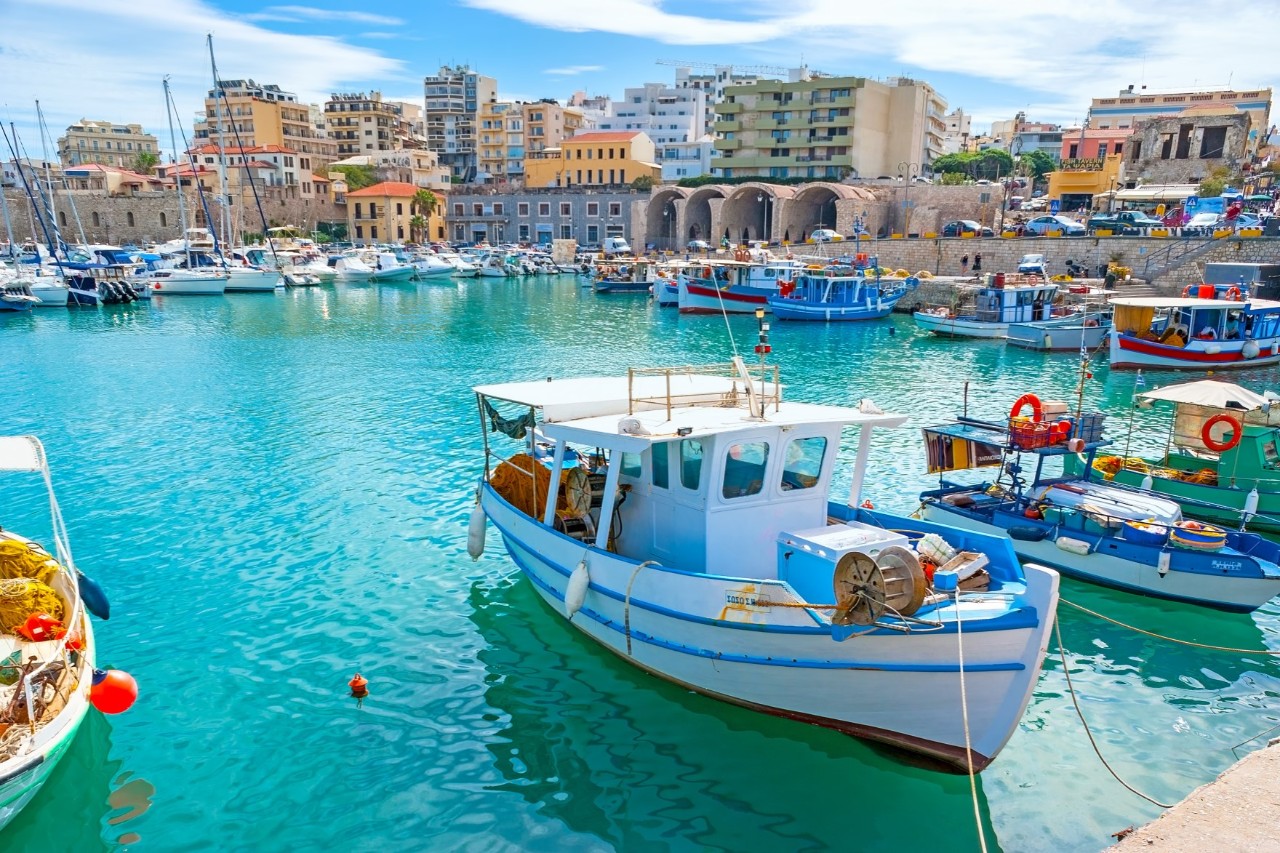 The picture shows a harbour basin with turquoise-blue water, in which there are many fishing boats all around. In the background, rows of houses can be seen.